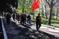 На фотографії колона людей іде парковою алеєю в сонячний день, де попереду крокує чоловік у камуфляжній формі з медалями на грудях, несучи великий червоний прапор та тримаючи червоні тюльпани.