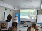 У світлій аудиторії жінка-спікер у білій сорочці виступає перед аудиторією на фоні великого екрана з презентацією «ПОВЕРНЕННЯ», поки поруч за столом із ноутбуком стоїть чоловік, а на передньому плані видно потилиці слухачів.