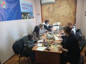 У кімнаті з великою старовинною картою та плакатами «Щастинський район» і «Луганщина — це Європа» група дітей та дорослих сидить за столом, беручи участь у майстер-класі, де інструкторка показує малюнок на фоні розкладених художніх матеріалів, напоїв та інформаційних буклетів.