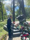На фотографії чоловік у темному одязі покладає букет квітів до підніжжя бронзового монумента у вигляді людської постаті на півсфері, оточеного численними кошиками та букетами червоних і білих квітів у сонячному зеленому парку
