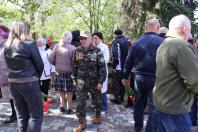 У центрі фотографії літній чоловік у військовій камуфляжній формі з численними медалями на грудях іде серед натовпу людей, які тримають червоні тюльпани біля меморіальної стіни в сонячний день.