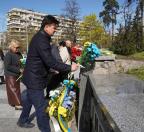 На фоні багатоповерхового будинку та весняних дерев чоловік у темній куртці покладає синьо-жовтий букет квітів до кам'яного меморіалу жертвам Чорнобиля, біля підніжжя якого вже стоять кошики з квітами та пам'ятними стрічками, а поруч чекають інші учасники церемонії
