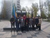На фотографії група з десяти людей різного віку з квітами в руках позує на фоні пам'ятника жертвам Чорнобиля та багатоповерхівки з великим патріотичним муралом Статуї Свободи