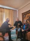 У кімнаті з жовтими фіранками та візерунчастим килимом на стіні двоє чоловіків сидять на дивані, посміхаються та тиснуть один одному руки біля двох великих упаковок підгузків для дорослих на підлозі, а поруч із ними сидить літня жінка.
