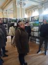 У великій світлій залі з класичними колонами група людей розглядає виставку дитячих малюнків на тему Чорнобиля, що розміщені на сітчастих стендах, при цьому на передньому плані стоїть жінка в коричневій куртці з бежевим рюкзаком, а за нею видно інших відвідувачів на фоні художніх робіт