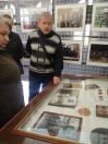 Чоловік у чорно-білому светрі з геометричним візерунком та жінка поруч уважно розглядають архівні фотографії та документи під склом горизонтальної вітрини, на фоні якої на сітчастих стендах розміщено інші світлини в рамках, що зображують пам'ятні заходи.