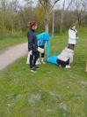 На цій світлині зображено чотирьох дівчат у куртках і рукавичках, які збирають сміття на зеленій парковій галявині біля ґрунтової стежки, пакуючи його у великі білі та блакитні поліетиленові мішки на фоні високих весняних дерев.