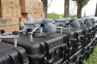 На передньому плані в ряд вишикувано кілька сучасних сірих дронів DJI Matrice 4T, кожен із яких стоїть на своєму міцному чорному захисному кейсі, а на задньому плані видно стоси картонних коробок та високі дерева на узліссі