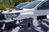 Два білі пікапи Toyota Hilux припарковані на фоні лісу, на їхніх капотах розкладено дрони та пульти дистанційного керування, а на капоті найближчого авто під обладнанням розгорнуто синьо-жовтий прапор України.