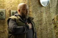 На фоні стіни, обтягнутої камуфляжною тканиною з розміщеними на ній почесними грамотами, лисий чоловік із густою сивіючою бородою у темно-сірому худі та оливковому жилеті стоїть у профіль, зосереджено приклавши праву руку до серця під час урочистого моменту