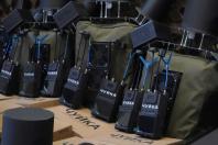 На передньому плані розташовані кілька портативних детекторів дронів чорного кольору з написом «ЧУЙКА» та антенами, які стоять на картонних коробках поруч із великими оливковими рюкзаками та складними технічними пристроями, оснащеними кулеподібними елементами та вентиляторами на тлі маскувальної сітки
