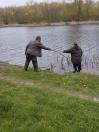 На березі водойми, вкритому зеленою травою з поодинокими жовтими кульбабами, чоловік у камуфляжних штанах і сірій куртці нахиляється та простягає довгу суху гілку жінці в темному одязі, яка стоїть біля самої води на фоні дерев із молодим весняним листям на протилежному боці.