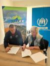Чоловік у худі з логотипами та світловолоса жінка у джинсовій жилетці сидять за дерев'яним столом на фоні банерів організацій «NEEKA» та «УВКБ ООН», де жінка підписує офіційні документи синьою ручкою.
