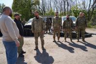 На відкритій сонячній ділянці біля дерев група з п’яти військовослужбовців у піксельному камуфляжі та двох чоловіків у цивільному одязі проводять зустріч, стоячи поруч із двома білими пікапами, біля яких на землі розставлено численні чорні захисні кейси з технічним обладнанням
