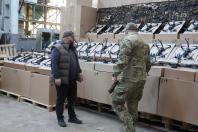 Всередині просторого ангару на фоні індустріальних конструкцій та маскувальної сітки чоловік у темному жилеті та військовослужбовець у камуфляжі стоять поруч із довгими рядами FPV-дронів, які акуратно розкладені на штабелях картонних коробок, підготовлених для відправки на передову.
