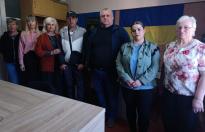 Група з семи осіб, серед яких жінка в червоному піджаку та чоловік у кепці з попередніх кадрів, стоять у ряд у приміщенні на фоні державного прапора України та темного стяга з військовою символікою, а на передньому плані видно частину масивного дерев’яного столу.