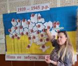 Біля патріотичного плаката з мапою України, прикрашеною червоними маками на тлі синьо-жовтого прапора, стоїть жінка в картатій сорочці, яка однією рукою прикріплює квітку до мапи, а в іншій тримає ще один паперовий мак, доповнюючи композицію з гаслом «Ніщо не забуте, Ніхто не забутий».