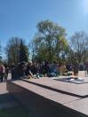 Група жінок у темному та повсякденному одязі нахилилася над гранітним постаментом. Вони обережно кладуть живі квіти (жовті та сині хризантеми, червоні гвоздики) до підніжжя меморіалу. Їхні обличчя зосереджені та серйозні.