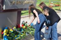 Дві молоді жінки та маленька дитина (хлопчик років 5-6) стоять біля підніжжя пам'ятника. Одна з жінок у чорній футболці та джинсах нахилилася, щоб обережно покласти квіти. Інша жінка у чорному піджаку стоїть поруч і притримує дитину за плечі. Хлопчик уважно спостерігає за процесом