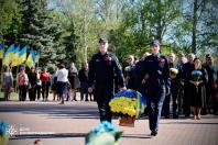 Попереду колони йдуть двоє молодих правоохоронців (хлопець та дівчина) у темно-синій формі та кепках. Разом вони несуть великий плетений кошик, наповнений жовтими та блакитними квітами, перев'язаний стрічкою національних кольорів. У кожного на грудях прикріплена червона квітка маку
