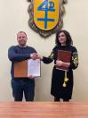 Чоловік у синьому лонгсліві та жінка в чорній сукні з жовтою вишивкою потискають одне одному руки, тримаючи в руках папки з документами на фоні світлої стіни з великим гербом синього та жовтого кольорів.