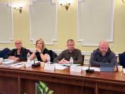 За довгим дерев'яним столом засідань на тлі світло-жовтої стіни з класичним білим оздобленням та бра сидять четверо людей, серед яких блондинка в окулярах і чорній куртці з ручкою, ще одна блондинка у чорній футболці, яка підперла підборіддя рукою, чоловік у коричневій сорочці та окулярах, а також бородатий чоловік у сірому худі, який дивиться в екран планшета перед собою, де перед кожним учасником розкладено документи, пляшки з водою, паперові стаканчики та встановлено настільні мікрофони й іменні таблички