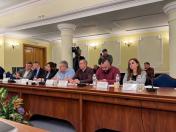 За довгим дерев'яним столом у залі засідань з масивними білими колонами та класичними настінними бра сидять семеро учасників заходу, серед яких жінки та чоловіки в офіційно-діловому та повсякденному одязі, що вивчають документи або дивляться вперед, перед якими встановлено мікрофони, іменні таблички та пляшки з водою, тоді як на задньому плані біля стіни стоять оператор із професійною відеокамерою та чоловік у бейсболці, який знімає подію на мобільний телефон