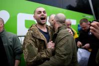 На світлині зафіксовано емоційний момент повернення з полону, де на тлі яскраво-зеленого пасажирського автобуса коротко стрижений український військовий у камуфляжній куртці з широко розкритим у радісному крику ротом міцно стискає руку та обіймає за плече свого побратима, повернутого спиною до кадру, в той час як навколо них стоять інші чоловіки у військовому та цивільному одязі