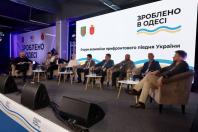 Група чоловіків сидить у м’яких кріслах на сцені під час офіційної панельної дискусії на тлі великого білого екрана з написом «Форум економіки прифронтового півдня України».