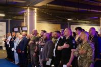 Велика група людей у цивільному одязі, костюмах та військовій формі стоїть у ряд у приміщенні, приклавши праву руку до серця під час урочистого моменту