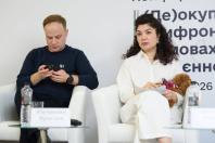 Чоловік у темному світшоті дивиться у телефон, а жінка у білому костюмі сидить поруч із маленьким рудим собакою на руках під час офіційного заходу на тлі білого банера з текстом.