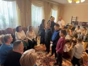 У просторій світлій кімнаті з великими вікнами та килимом на підлозі зібралася велика група дорослих і дітей, де частина людей сидить на дивані вздовж стіни, а діти стоять і роздивляються коробки з подарунками в центрі приміщення.