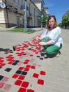 Жінка у білій сорочці з зеленою вишивкою та яскравих зелених штанах присіла на пішохідній вулиці міста, малюючи пензлем червоно-чорний орнамент вишиванки безпосередньо на тротуарній бруківці.