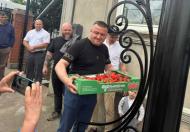 Чоловік у чорній футболці тримає в руках великий зелений картонний лоток, наповнений стиглою червоною полуницею, стоячи на вулиці біля кованих воріт в оточенні інших людей, поки хтось на передньому плані знімає його на мобільний телефон.