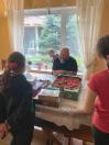 Лисий бородатий чоловік у чорній футболці посміхається, сидячи біля вікна за столом, на якому стоїть великий зелений лоток зі стиглою полуницею та коробки солодощів, а перед ним спиною до кадру стоять двоє дітей.