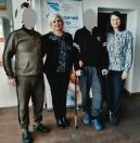 Дві жінки та двоє чоловіків, один із яких спирається на милиці, стоять у ряд та позують для спільного фото в приміщенні на тлі інформаційного банера з логотипом.
