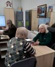 У світлому офісному кабінеті біля великого дзеркала білява жінка в зеленому піджаку спілкується з відвідувачкою за дерев'яним столом, поки на задньому плані інший чоловік працює за комп'ютером.