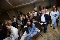 Люди в офіційному та повсякденному вбранні сидять рядами на стільцях у конференц-залі під час заходу, тримаючи в руках папери або телефони.