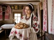 Молода жінка в українському традиційному костюмі з вишитими червоно-чорними квітами рукавами тримає на вишитому рушнику круглий святковий коровай, оздоблений виліпленими з тіста квітами та колосками, на фоні автентичного інтер'єру старовинної хати