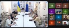 Скриншот онлайн-наради за участю Луганської ОВА, де ліворуч у великому вікні показано залу засідань із сімома посадовцями за довгим столом на тлі державних прапорів та банера «official media center», а праворуч — сітку з дванадцятьма віконцями інших учасників відеоконференції