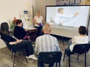 Блондинка у білій мереживній блузі проводить презентацію в кабінеті, тримаючи фіолетову папку біля великого екрана проєктора з трансляцією телепередачі «Сніданок з 1+1», у той час як четверо слухачів сидять на стільцях півколом і дивляться на екран
