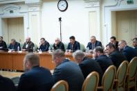 Численні учасники офіційного засідання в ділових костюмах сидять двома рядами за довгими дерев'яними столами у світлій залі з класичною архітектурою та круглим годинником на стіні