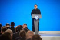 Президент України Володимир Зеленський у чорній сорочці виступає за сірою трибуною перед аудиторією на тлі яскраво-блакитного екрана