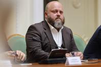 Портрет великим планом лисого чоловіка з густою сивіючою бородою в коричневому піджаку та білій сорочці, який сидить за столом засідань перед мікрофоном під час офіційного заходу.