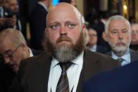 Портрет великим планом лисого чоловіка з густою русявою бородою у коричневому костюмі та краватці, який зосереджено дивиться повз камеру на тлі інших людей розмитого заднього плану
