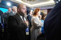 Лисий чоловік із густою бородою в коричневому костюмі та рудоволоса жінка у білій вишиванці стоять у залі серед інших людей, притиснувши праві руки до серця під час офіційного заходу