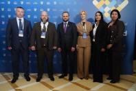Група з трьох чоловіків та трьох жінок у діловому вбранні стоїть у ряд для спільного фото на тлі синього брендованого банера з логотипами офіційного заходу.
