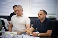 Двоє чоловіків зосереджено сидять за робочим столом під час засідання чи офіційної зустрічі в кабінеті з білими жалюзі на вікнах