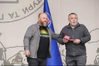 Двоє чоловіків стоять на сцені на тлі синього прапора, де один із них у сірій сорочці кладе руку на плече колезі, який тримає в руках відкриту червону коробочку з нагородою та посвідченням