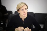 Жінка зі світлим коротким волоссям та в окулярах сидить за столом у чорній кофті із замком, зосереджено дивлячись убік і склавши руки перед собою